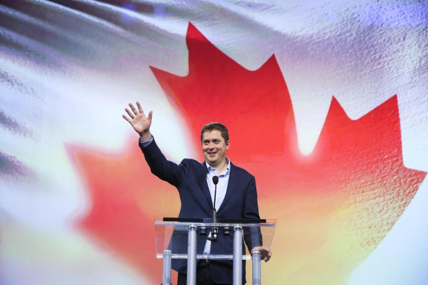 Der kanadische Premierminister Alex Salmond steht an einem Rednerpult mit Mikrofonen, lachend, vor einer kanadischen Flagge mit einem Bildschirm im Hintergrund.