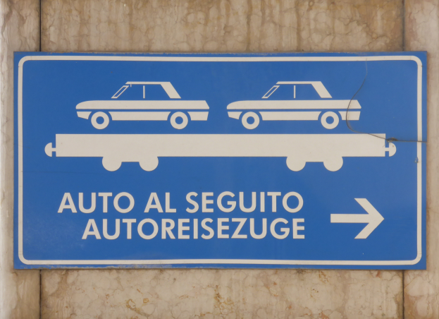 Ein blaues Schild an einem Gebäude mit der Aufschrift "Auto al Seguito Autoreisezuge" und zwei Autofotos.