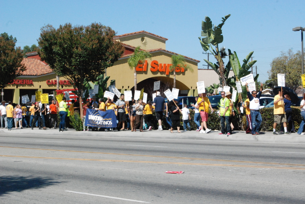 Eine Gruppe von Menschen mit Schildern und einer Baumreihe, Büschen, einem Banner, einem Gebäude mit Namensschildern, Autos und einer Wahllokal im Hintergrund.