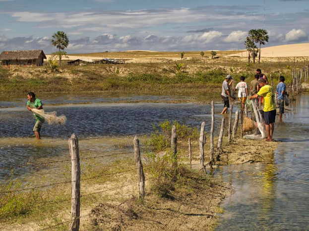 Eine Gruppe von Menschen, die in flachem Wasser stehen und gehen, Fische fangen, mit einem hölzernen Zaun in der Mitte, Boden und Wasser unten, Himmel oben und Bäume mit Hütten im Hintergrund.