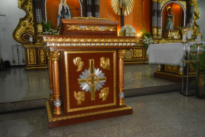 Ein Altar in einer Kirche mit einem Kreuz, Statuen, Blumenbouquets, Kerzen, einem tisch mit einer Decke und anderen Gegenständen, wobei die Wände mit Lichtern geschmückt sind.