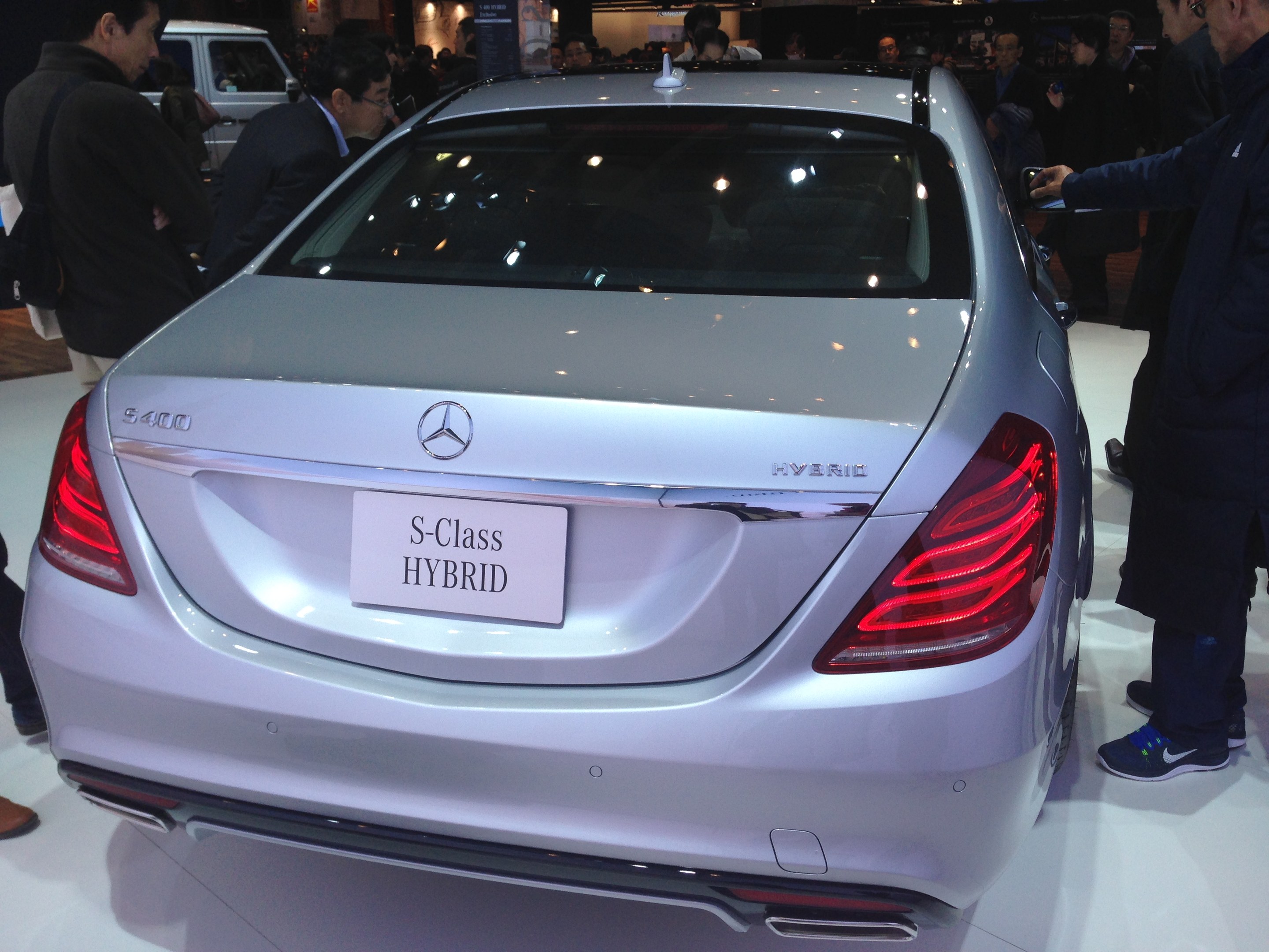 Silberner Mercedes S-Klasse Hybrid auf einer Automesse zu sehen, umgeben von Menschen in bunten Kleidern, mit anderen Fahrzeugen und Beleuchtung im Hintergrund.