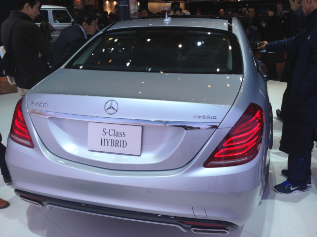 Silberner Mercedes S-Klasse Hybrid auf einer Automesse zu sehen, umgeben von Menschen in bunten Kleidern, mit anderen Fahrzeugen und Beleuchtung im Hintergrund.