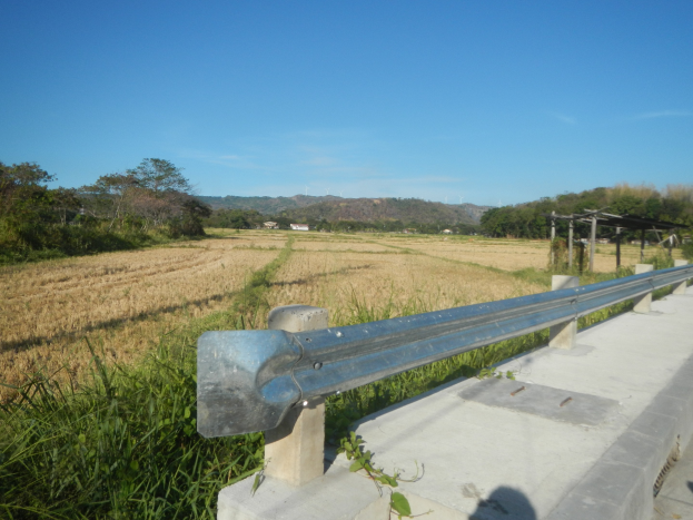 Eine Straße mit einer Metallschutzplanke, grünem Gras, Bäumen, Hügeln und einem hellblauen Himmel.