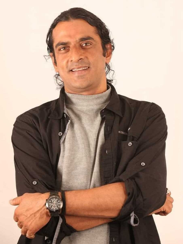 Ein Mann in einem schwarzen und aschfarbenen Kleid mit verschr├Ąnkten Armen, der eine Uhr tr├Ągt und vor einem wei├čen Hintergrund steht.