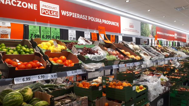 Ladeninnere mit verschiedenen Früchten und Gemüsen in Kartons mit Preisschildern, Werbetafeln an den Wänden und Deckenbeleuchtung.