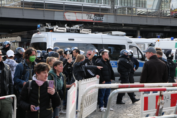 Eine Gruppe von Demonstranten steht hinter Barrikaden vor Polizeifahrzeugen, einige tragen Helme und filmen mit Handys, mit einer Brücke und Gebäuden im Hintergrund während eines Auflösungsmanövers in Berlin.