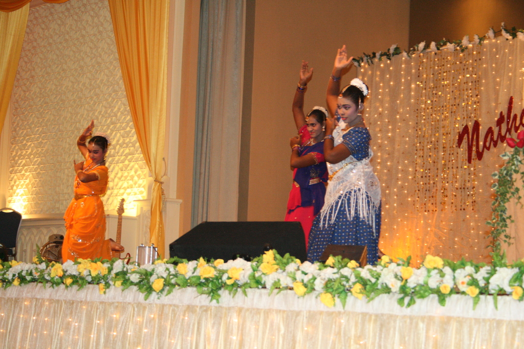 Eine Bühne mit weißen und gelben Blumen, drei tanzenden Frauen, einem schwarzen Objekt, einem cremefarbenen Vorhang, Lichtern, einer braunen Wand und anderen Gegenständen im Hintergrund.