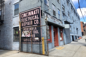 Außenansicht des Cincinnati Electrical Repair Co.-Gebäudes mit Fenstern, Türen, einem Schild, einem Metallzaun, einem Müllcontainer, Strommasten, Drähten, Bäumen und einem bewölkten Himmel.