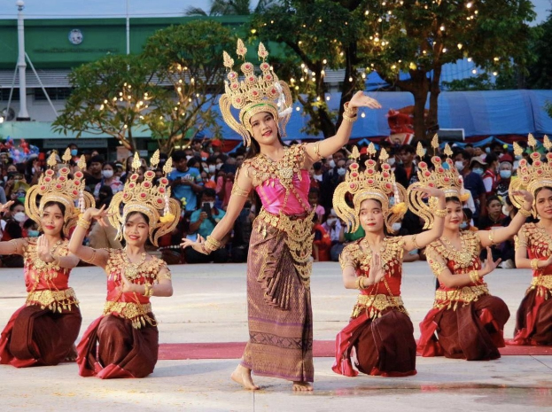 Gruppe von Frauen in traditioneller Thai-Kleidung, die einen Tanz vor einem sitzenden und stehenden Publikum im Freien aufführen, mit Bäumen, Lichtern, Pfosten, einem Gebäude und einem klaren blauen Himmel im Hintergrund.