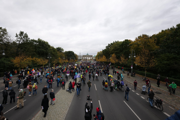 Große Gruppe von Menschen marschiert durch eine baumbestandene Straße in Berlin, einige halten Kameras, mit einem Gebäude und wolkenlosem Himmel im Hintergrund.