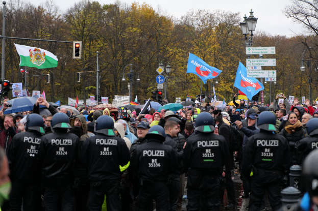 Eine große Gruppe von Polizisten steht vor einer Menge mit Fahnen und Schirmen, im Hintergrund sind Laternenmasten, Verkehrsampeln, Verkehrszeichen, Bäume und ein klarer Himmel zu sehen.