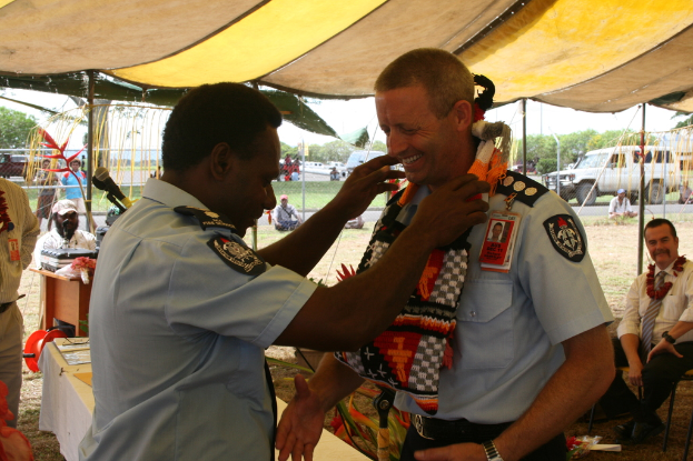 Zwei Beamte in einheitlicher Uniform, einer sitzt auf einem Stuhl und der andere steht in der Nähe eines Tisches mit Papieren, Kartons und einem Koffer; ein Mann in einem weißen Kleid sitzt neben dem Tisch; Menschen stehen und gehen auf dem Rasen; Fahrzeuge auf der rechten Seite; ein Zelt oben; Bäume, Pflanzen, Gras und Himmel im Hintergrund.