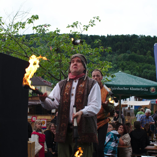 Eine Person hält Feuer in der Mitte einer Versammlung von Zelten und Ständen, mit Bäumen und Himmel im Hintergrund.