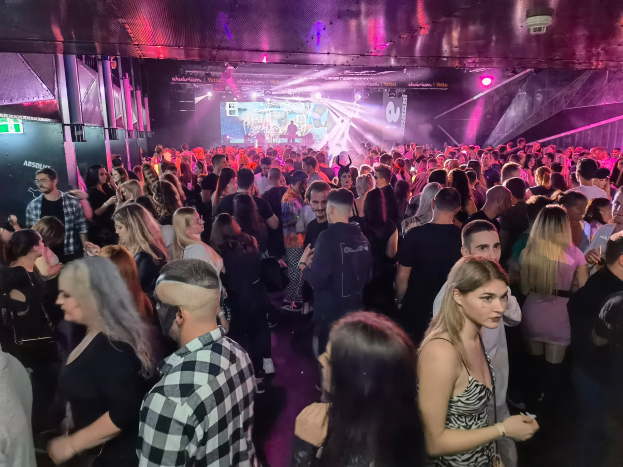 Eine große Gruppe von Menschen in einer Diskothek, beleuchtet von Deckenlichtern und umgeben von Säulen, mit einem Bildschirm im Hintergrund, der auf ein Absolut Nightlife-Event hindeutet.