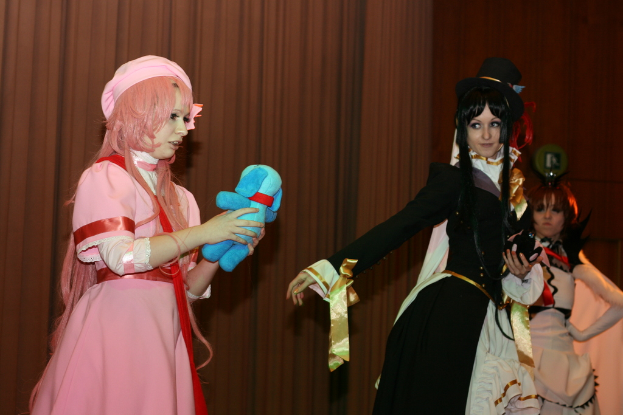 Drei Frauen führen ein Drama auf, wobei eine in einem rosafarbenen Kleid mit einem blauen Teddybär und eine andere in einem schwarzen Kleid nach links schaut, während ein brauner Vorhang im Hintergrund zu sehen ist.