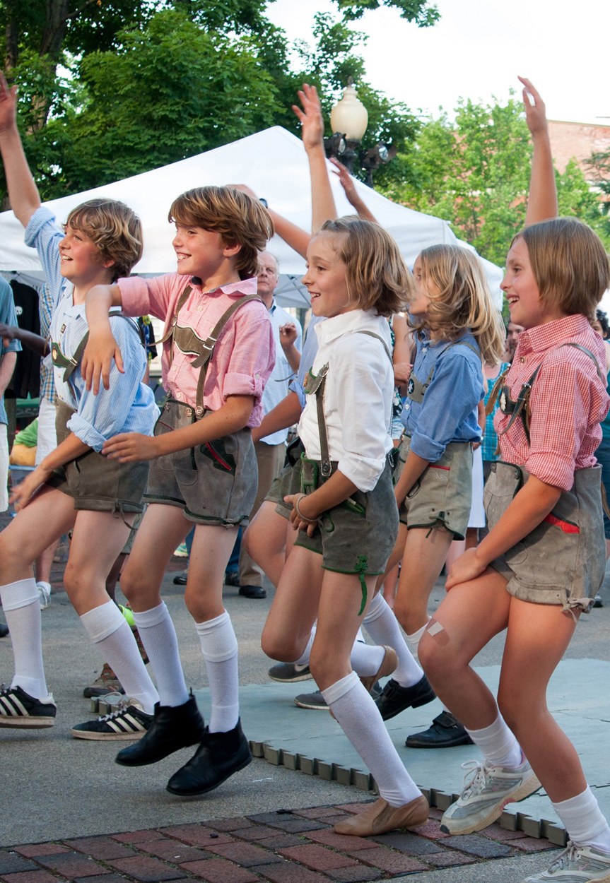 Kinder in traditioneller bayrischer Tracht auf einer Straße tanzend, umgeben von einer Menge, Zelten, Laternenmasten, Bäumen, Gebäuden und einem klaren blauen Himmel.