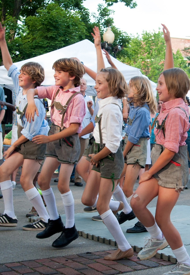 Kinder in traditioneller bayrischer Tracht auf einer Straße tanzend, umgeben von einer Menge, Zelten, Laternenmasten, Bäumen, Gebäuden und einem klaren blauen Himmel.