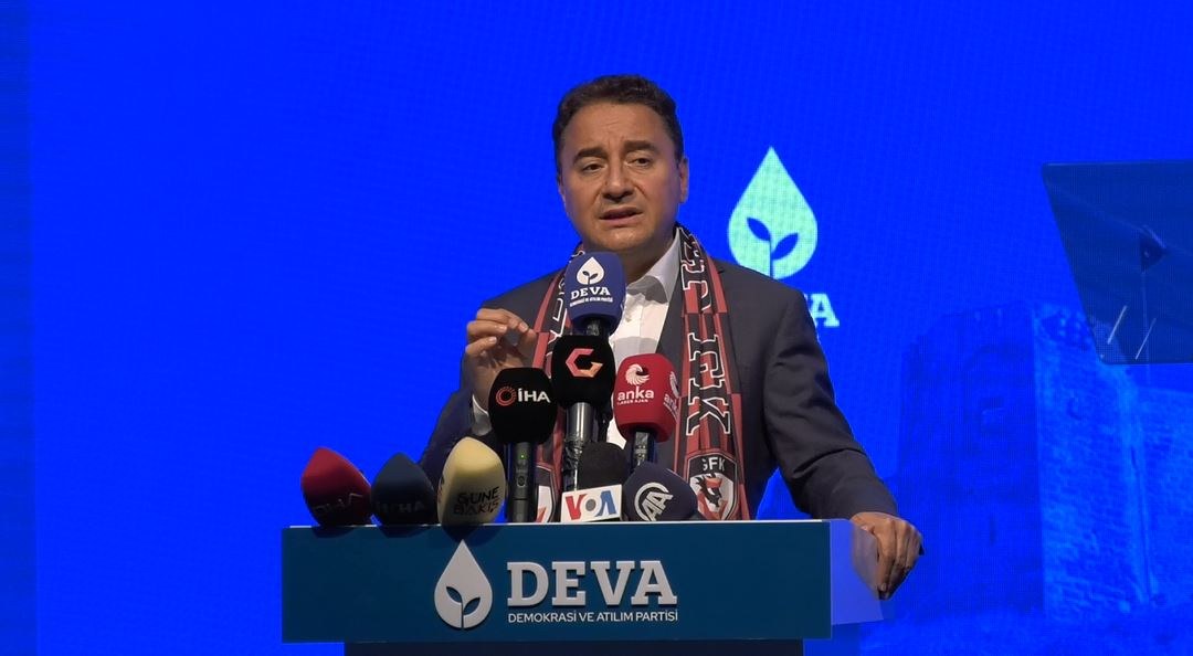 Mann in Mantel und Schal steht an Pult mit Mikrofonen, spricht zu Publikum; Pultschild reads 'deva ceo sagt, es ist keine gute Idee für die Zukunft'; Bildschirm und Objekt sind im Hintergrund sichtbar.