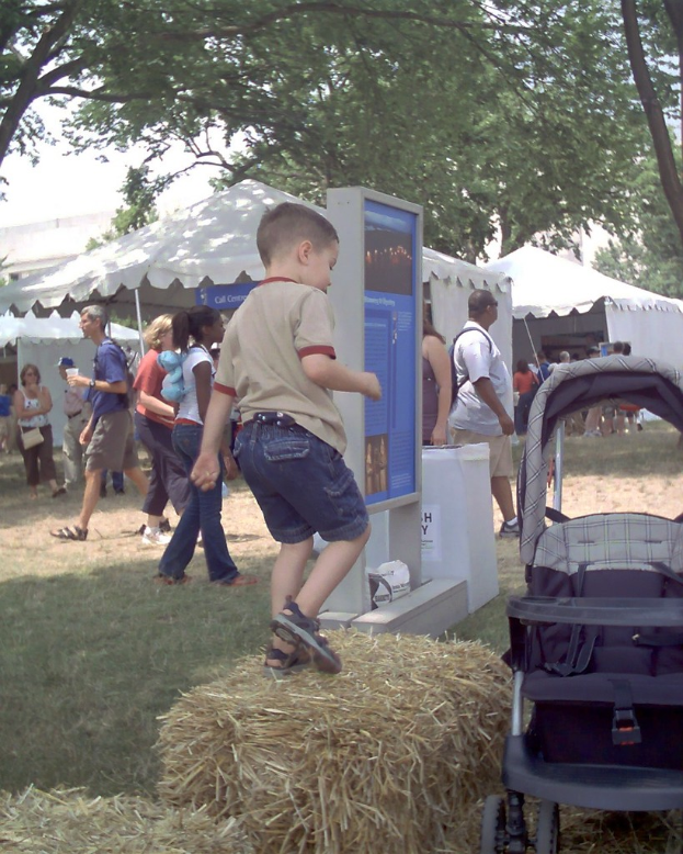 Eine Szene mit mehreren Zelten und Menschen, die drumherum gehen, ein Anzeigebrett vorne, trockenes Gras mit einem spielenden Kind und Bäume auf beiden Seiten.