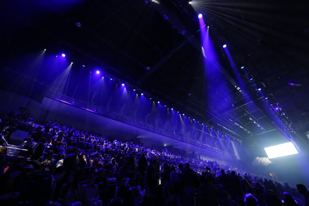 Große Menge sitzt vor einer Bühne mit Deckenbeleuchtung und einem Bildschirm auf der rechten Seite.