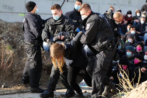 Gruppe maskierter Personen mit Mützen, einige bewaffnet, umringt eine Frau, die von der Polizei in einer städtischen Umgebung mit Pflanzen, Steinen und einer protestbezogenen Wandtexttafel eskortiert wird.