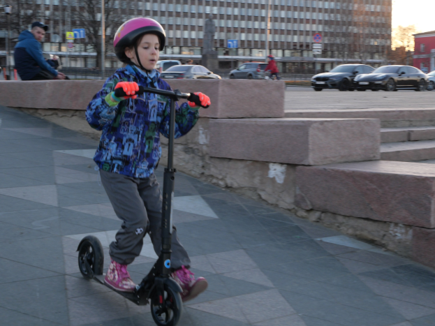 Ein junger Junge in einem Helm und Handschuhen fährt auf einem Roller eine Treppe auf dem Gehweg hinunter, mit Fahrzeugen, Menschen, Bäumen, Polen, Brettern, Gebäuden und einem klaren blauen Himmel im Hintergrund.