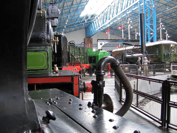 Mehrere Zugmaschinen sind in einem Go-down mit zahlreichen Eisenstäben an der Oberseite geparkt. Rechts verläuft ein Zaun entlang einer Eisenbahnschiene mit einem Zug. Zwei Individuen stehen in der Mitte der Szene in der Nähe des Zauns.