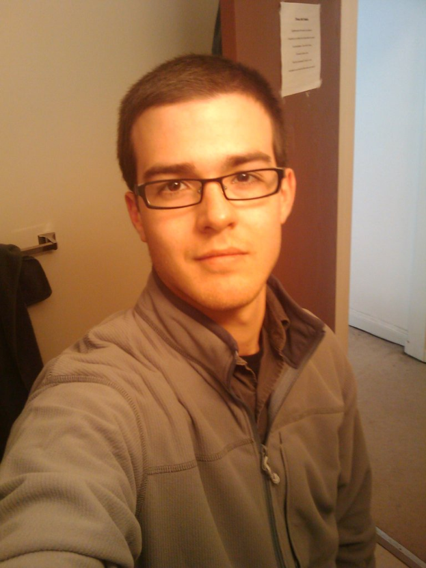 Ein Mann in einem Wams und Brillengläsern steht lächelnd da, mit einem Blatt Papier an der Wand hinter ihm und einem Tuch, das an einem Bügel zu seiner Linken hängt.