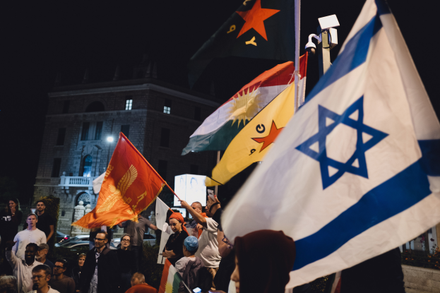Eine Gruppe von Menschen steht vor einem Gebäude und hält Fahnen während einer anti-israelischen Demonstration in der Nacht, mit Fahrzeugen und Pflanzen im Hintergrund.