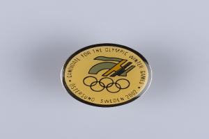 Ein runder Abzeichen mit schwarzem Hintergrund und goldfarbenem Olympische Winterspiele-Logo in der Mitte, mit der Aufschrift "Olympische Winterspiele" in fetter weißer Schrift.