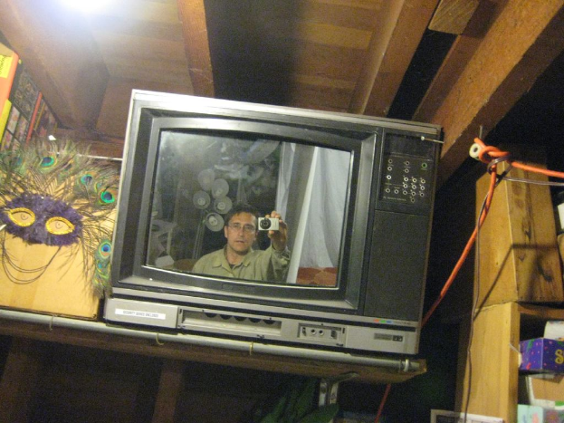 Ein Fernseher mit einer Person umgeben von Pfauenfedern, Büchern und Kisten.