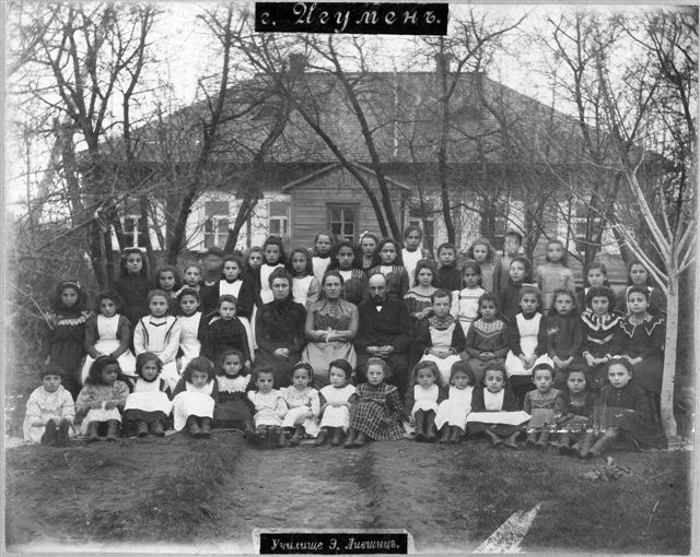 Schwarzes und weißes Foto einer Gruppe von Kindern, die vor einem Fenster mit einem Gebäude posieren, umgeben von Bäumen und einem klaren Himmel, mit Text unten, der lautet: "Jugoslawien, die erste Schule der Welt."