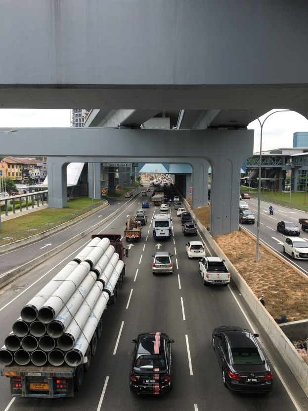 Eine vielbefahrene Autobahn mit mehreren Fahrzeugen, einer Brücke darüber, Straßenlaternen, Gras, Gebäuden, Bäumen und einem klaren Himmel.