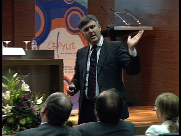 Ein Mann in einem schwarzen Blazer steht an einem Podium mit Mikrofonen, spricht in eines hinein und hat ein Blumentopf oder einen Blumenstrauß daneben. Vor ihm sitzen zwei Männer und eine Frau auf Stühlen. Hinter ihm ist eine braune Wand und eine bunte Tafel mit Text zu sehen, was auf eine Tagungsraum-Situation hindeutet.