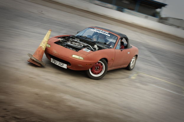 Eine Person, die ein rotes Auto auf einer Rennstrecke fährt, mit einem Verkehrskegel vor sich und einem unscharfen Hintergrund, der hohe Geschwindigkeit andeutet.