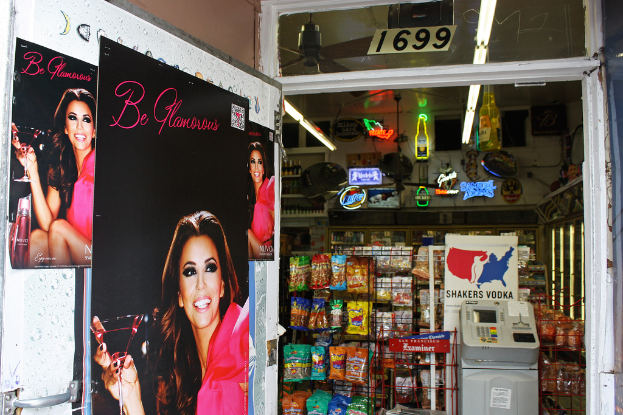 Eine Innenansicht mit einer Tür mit Plakaten einer Frau links, einem Geschäft mit verschiedenen Artikeln in der Mitte, einem Geldautomaten und einer Texttafel rechts sowie einer Nummerntafel oben.