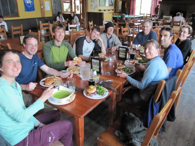 Eine Gruppe von Menschen sitzt um einen Tisch mit Tellern, Schüsseln, Gläsern, Flaschen, einem Wasserglas, Karten, einem Tuch und Essen, lachend, mit einer Jacke auf einem der Stühle, während einige weitere Personen an anderen Tischen sitzen, mit einem Banner, Bildern und Postern an der Wand und einem Fenster mit Vorhängen.