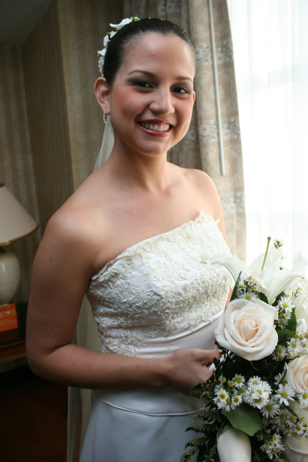 Eine Braut steht lachend in der Mitte, hält einen Strauß und hat einen Vorhang und eine Wand im Hintergrund sowie eine Lampe auf einem Tisch links.