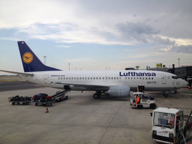 Ein Lufthansa Airbus A320-200 steht auf dem Frankfurter Flughafen geparkt, umgeben von diversen Bodenfahrzeugen, Wagen, Verkehrskegeln, Polen und Texttafeln unter einem bewölkten Himmel.