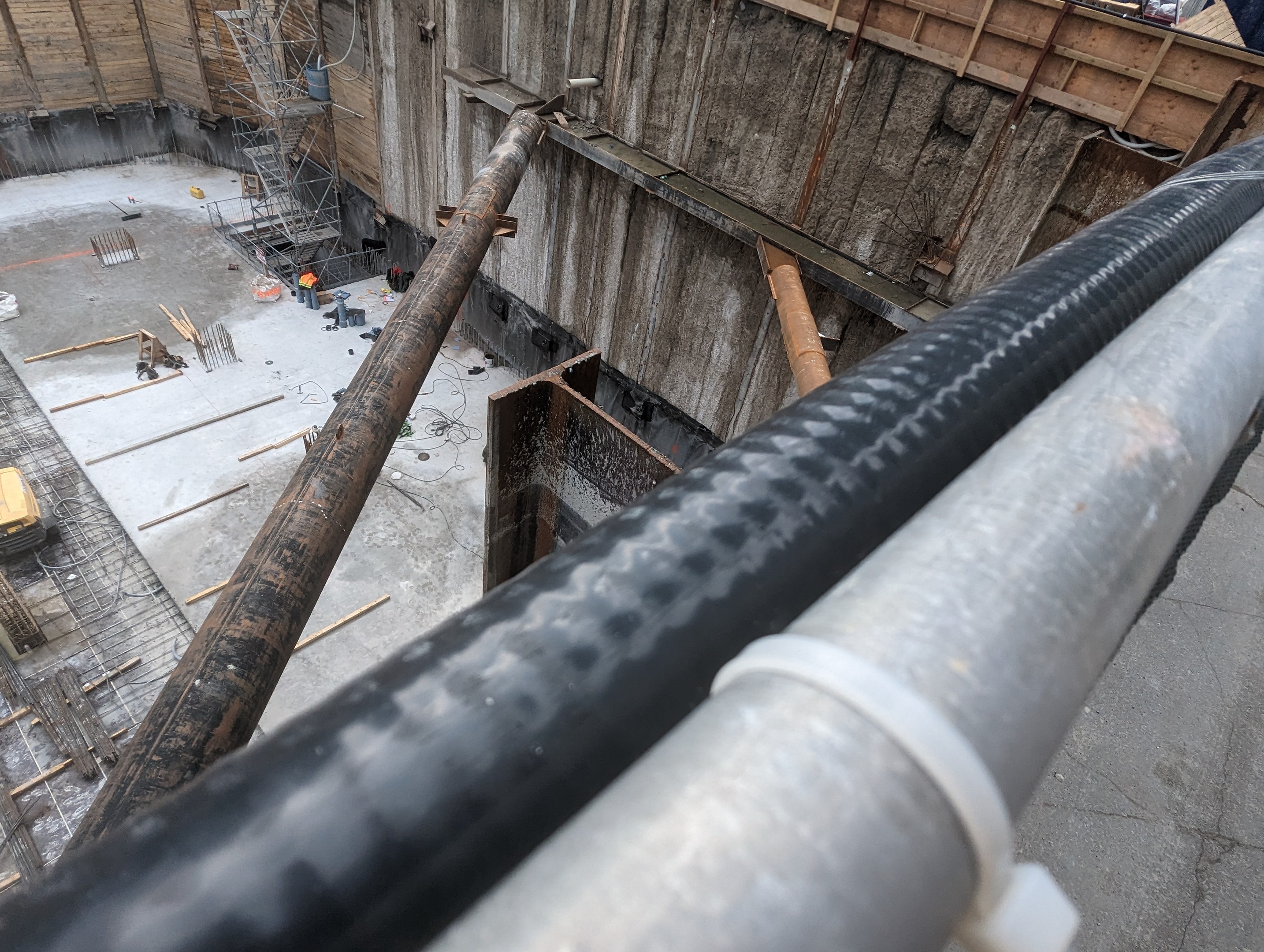 Großes Metallrohr auf einer Baustelle umgeben von Arbeitern, Holzwerkstoffen und Equipment, mit einer Person in einem blauen Hemd auf der rechten Seite und Fahrzeugen und einem Gebäude im Hintergrund.