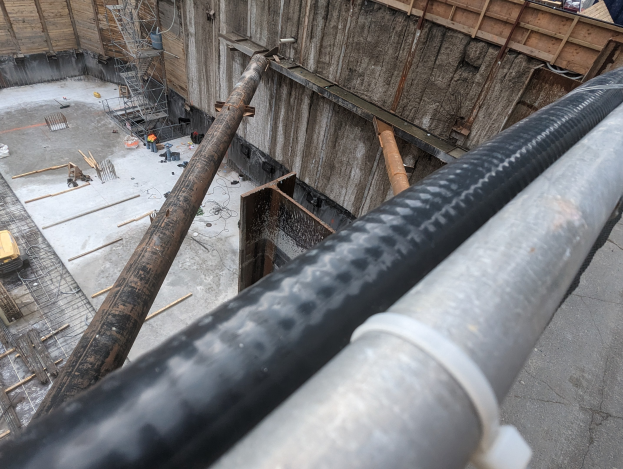 Großes Metallrohr auf einer Baustelle umgeben von Arbeitern, Holzwerkstoffen und Equipment, mit einer Person in einem blauen Hemd auf der rechten Seite und Fahrzeugen und einem Gebäude im Hintergrund.