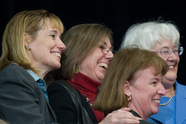 Mehrere ältere Frauen stehen im Vordergrund, lächeln und schauen nach rechts, mit einem schwarzen Hintergrund dahinter.