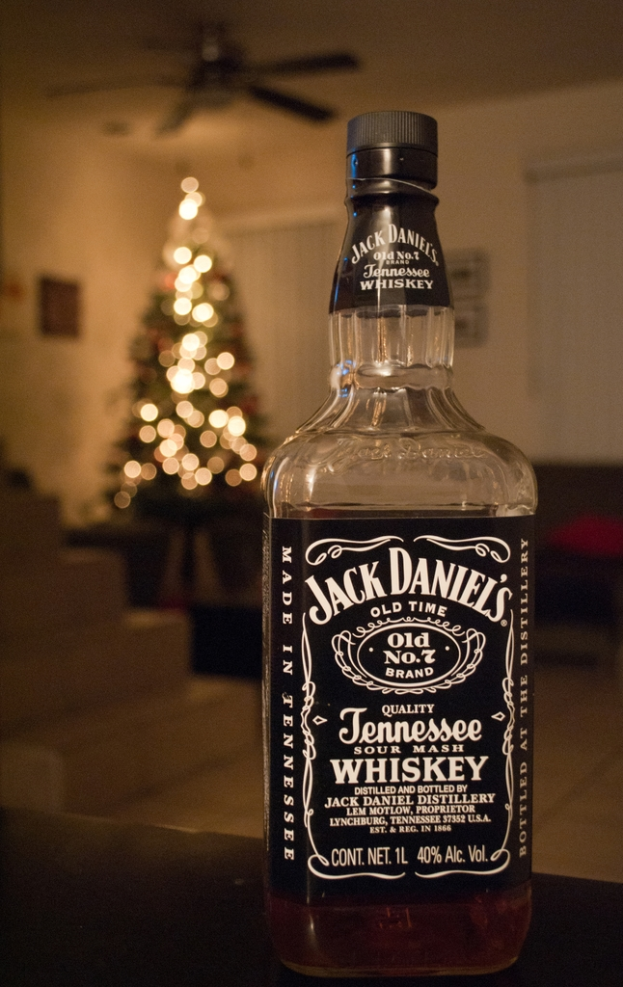 Eine Glasflasche mit der Aufschrift "Jack Daniel's" steht auf einem Tisch vor einem beleuchteten Weihnachtsbaum, mit einem Ventilator darüber und einer weißen Wand im Hintergrund.