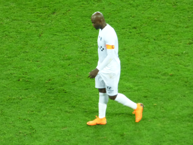 Ein Fußballspieler in einem weißen T-Shirt, weißen Shorts und orangefarbenen Schuhen, der über ein saftiges grünes Feld läuft.
