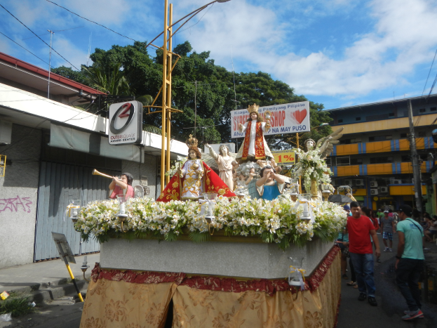 Ein Paradewagen mit Statuen von Menschen, Blumen und anderen Gegenständen, begleitet von Texttafeln, Laternenmasten, Kabeln, Gebäuden, Bäumen und einem bewölkten Himmel im Hintergrund.