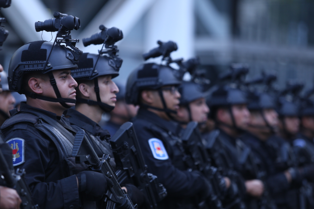 Eine Gruppe von Polizisten in schwarzen Uniformen und Helmen, die Waffen halten, steht zusammen in einer unscharfen militärischen Umgebung.