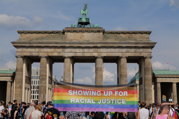 Eine Gruppe von Menschen, die eine "Racial Justice"-Schleife vor dem Brandenburger Tor in Berlin, Deutschland, halten, mit den Säulen und der Statue des Tors im Hintergrund und Gebäuden und einem bewölkten Himmel.