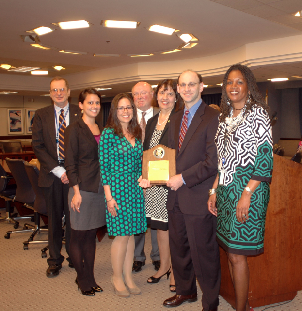 Eine Gruppe von Menschen, die in der Mitte stehen und einen Pokal halten, mit Stühlen und Porträts links, einem Schreibtisch rechts und Lampen oben auf dem Bild.