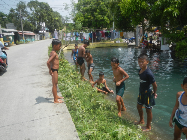 Kinder spielen in flachem Straßenwasser, einige stehen, andere sitzen auf dem Gras; eine Person sitzt auf einem Motorrad links, mit Häusern, Bäumen, Strommasten und Drähten im Hintergrund unter einem klaren blauen Himmel.